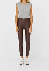 Vrouw in een mouwloos grijs top met de tekst "Say Less Do More", bruine hoog-heid leren broek en witte sneakers, rechtopstaand.
