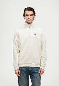 Jasno beżowy sweatshirt z półzamkiem, wykonany z miękkiego polaru, z paskiem na klatce piersiowej, małym detalem logo oraz ściągaczami przy mankietach i hemie.