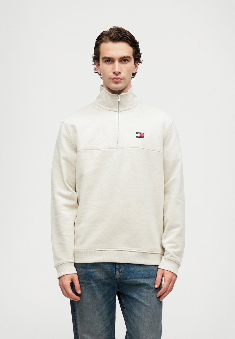 Jasno beżowy sweatshirt z półzamkiem, wykonany z miękkiego polaru, z paskiem na klatce piersiowej, małym detalem logo oraz ściągaczami przy mankietach i hemie.