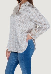 Chemise à manches longues à boutons en tissu crème avec un motif géométrique beige et marron. Dotée d'un col classique et de poignets à motifs.