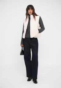 Gilet trapuntato rosa chiaro indossato su un dolcevita grigio scuro, abbinato a jeans larghi scuri. Una borsa nera e calzature completano il look.