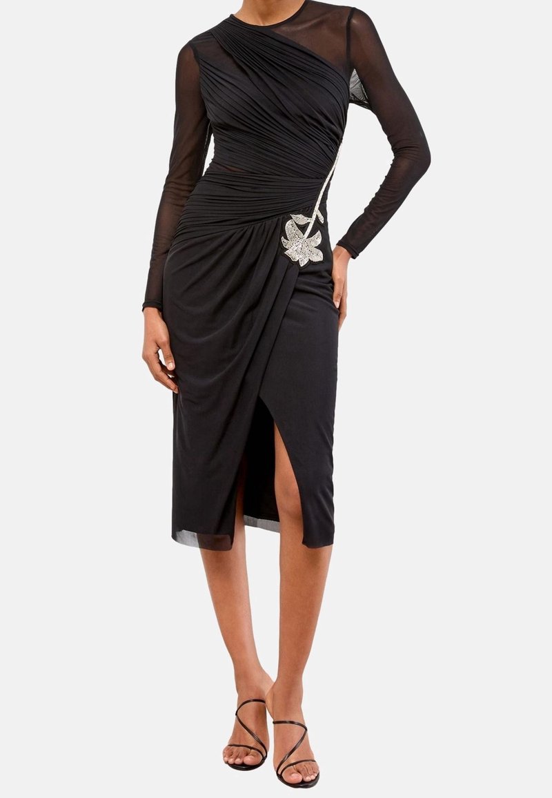 Femme portant une robe midi noire avec des manches longues transparentes, des détails froncés, une fente à l'avant et une décoration florale argentée à la taille.