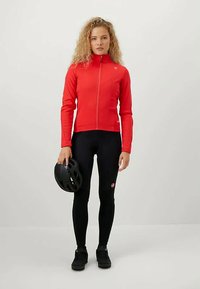 Červená bunda na zips, čierne legíny a čierne topánky. Drží čiernu prilbu na bicykel. Hladká textúra, priliehavý dizajn a logo na bunde.