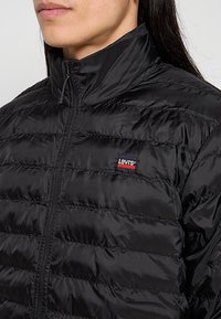 Gros plan sur une personne portant une veste noire matelassée Levi's avec une fermeture éclair à l'avant et un col montant, montrant le logo Levi's sur la poitrine.