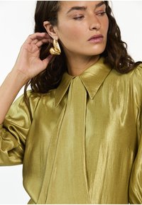 Glanzend olijfgroen blouse met een geplooide kraag en gestructureerde schouders, voorzien van een centrale plooi. Gouden statement oorbellen voegen visuele interesse toe.