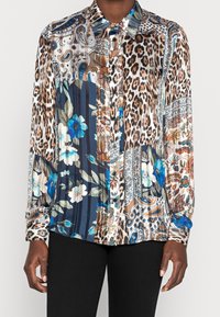 Blouse à manches longues avec boutons, présentant des motifs mixtes incluant des taches de léopard, des fleurs bleues et des motifs paisley dans des tons marron, bleu et blanc.