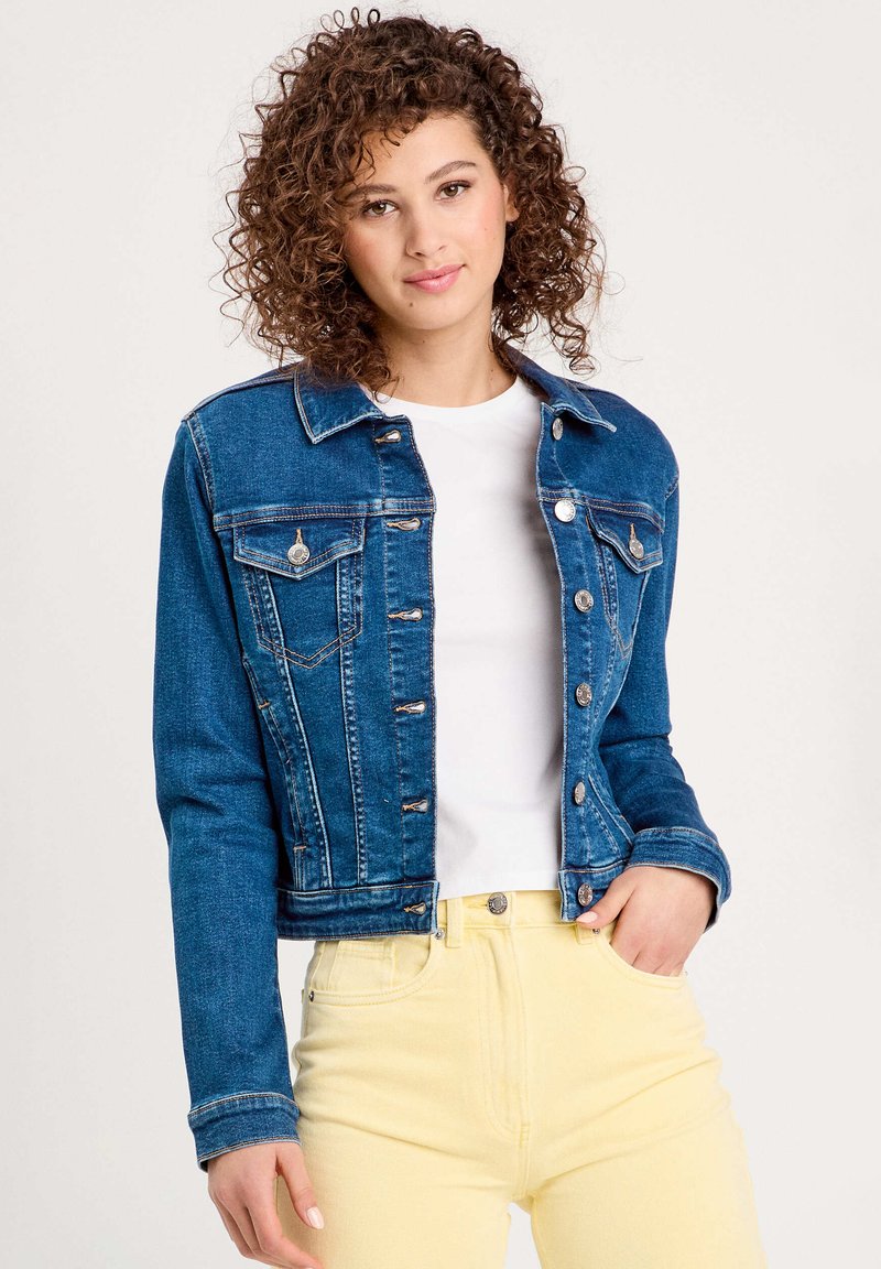 Denim jacket with a dark blue wash, classic collar, front pockets, silver button hardware, fitted shape, worn over a white shirt and yellow pants.