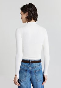 Frau mit kurzen lockigen Haaren, die ein weißes, geripptes Langarmoberteil und hochgeschnittene blaue Jeans mit einem dunkelbraunen Gürtel trägt, von hinten gesehen.