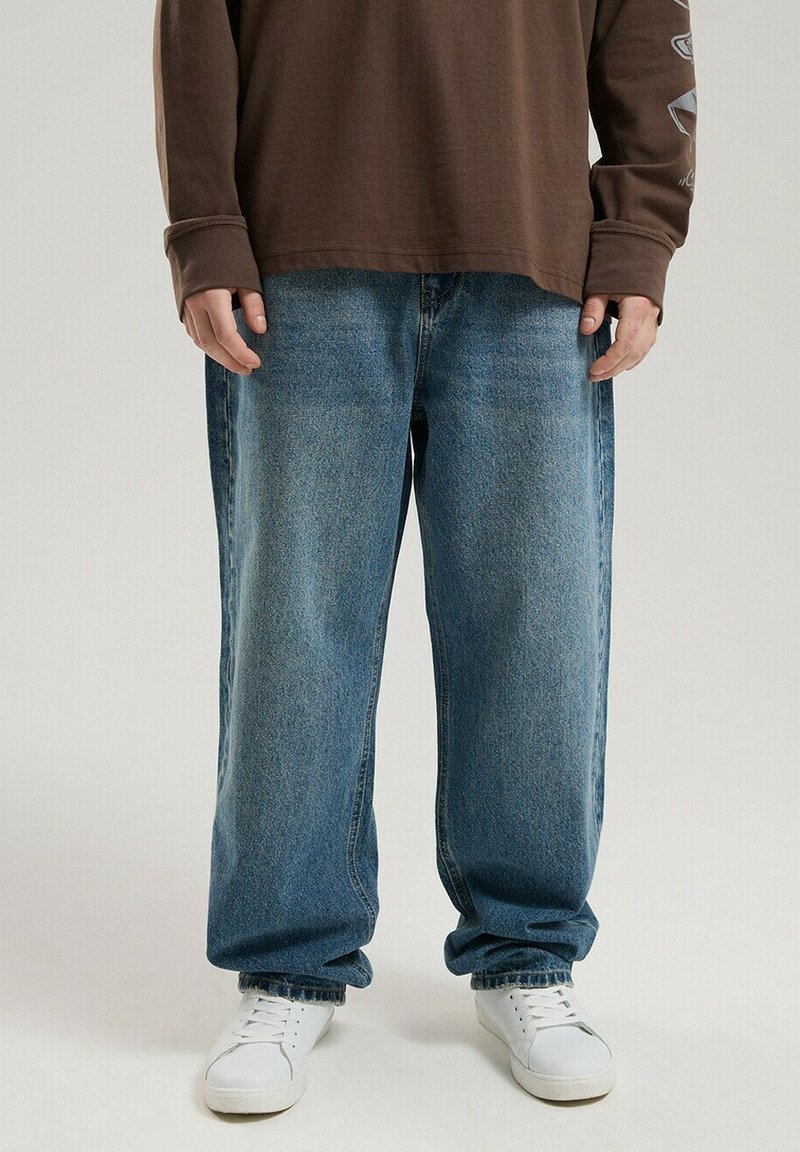 Niebieskie dżinsy o luźnym kroju z delikatnym efektem sprania i prostymi nogawkami. Noszone z brązową marynarką z długim rękawem i białymi sneakersami.