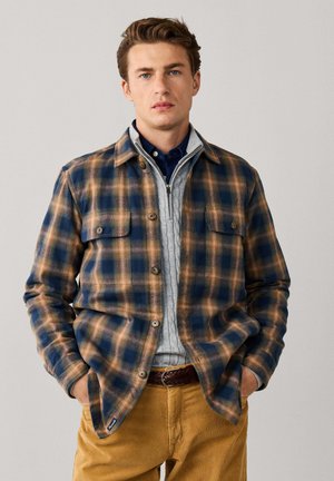 Joven con una chaqueta de cuadros azul y marrón sobre un jersey gris con cremallera y pantalones mostaza, de pie con las manos en los bolsillos.