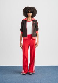 Athletic outfit featuring a brown zip-up jacket with red accents and logo, paired with red track pants with white stripes. Smooth texture.