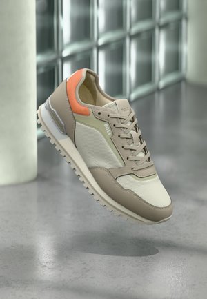 Baskets beige et blanches avec accent orange au talon, semelle texturée et design à lacets, suspendues au-dessus d'un sol gris près d'un mur en blocs de verre.