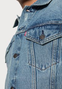 Närbild av ett blått denimjacka bröstficka med knapp och röd Levi's-märke, bärs av en person som står något vänd bort.