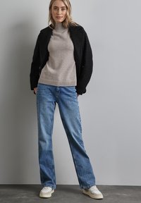 Černá strukturovaná bunda, béžový žebrovaný rolák, světlé modré džíny a bílé tenisky. Oblečení má uvolněný střih a vrstvený vzhled.