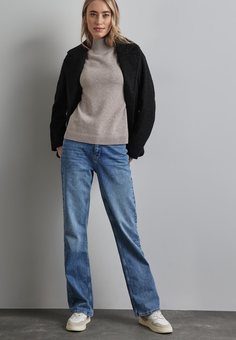 Černá strukturovaná bunda, béžový žebrovaný rolák, světlé modré džíny a bílé tenisky. Oblečení má uvolněný střih a vrstvený vzhled.