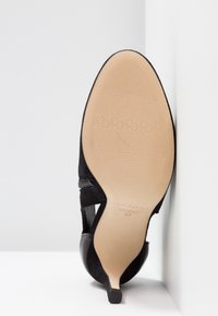 Chaussure à talon aiguille en daim noir, dotée d'un bout pointu, de découpes sur les côtés et d'un talon élancé. La semelle est beige avec un motif texturé.