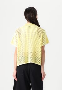 Top giallo a maniche corte, lavorato a maglia, con un design leggero e trasparente, dotato di colletto e orlo dritto, indossato sopra pantaloni scuri.