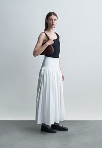 Žena stojaca v čiernom tielku bez rukávov, dlhej bielej sukni, čiernych topánkach, sivých ponožkách, drží malú hnedú kabelku cez plece.