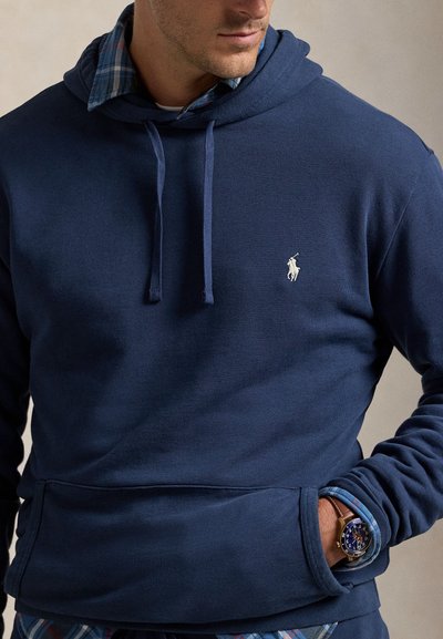 Navyblå hættetrøje med kængurulomme, ribbede manchetter og broderet logo på brystet. Båret over en ternet skjorte med krave.