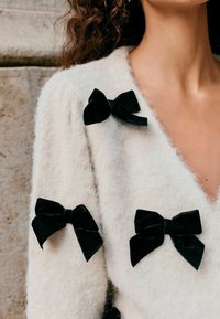Cremfarbener Cardigan aus fuzzy Stoff mit mehreren schwarzen Samtbändern, die vorne befestigt sind. V-Ausschnitt-Design mit kurzen Ärmeln.