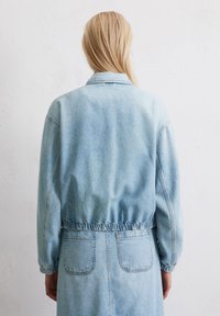 Eine Person mit langen blonden Haaren trägt eine hellblaue Jeansjacke und wird von hinten vor einer schlichten weißen Wand gesehen.