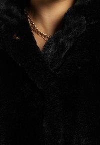 Sort faux fur frakke med en blød, fyldig tekstur, der har en spidse krave og en metallisk detalje. Nærbillede viser stofdetaljerne.