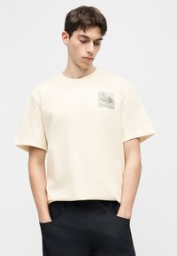 Νέος άνδρας με καστανά μαλλιά που φοράει μπεζ μπλουζάκι The North Face και μαύρο παντελόνι, με τα χέρια στις τσέπες, κοιτάζοντας προς τα κάτω αριστερά του.