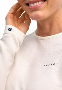Maglietta a maniche lunghe color crema realizzata in tessuto strutturato, con colletto a costine e logo stampato "FALKE" vicino al fondo. Accentuata da una "R" scura sul polsino.