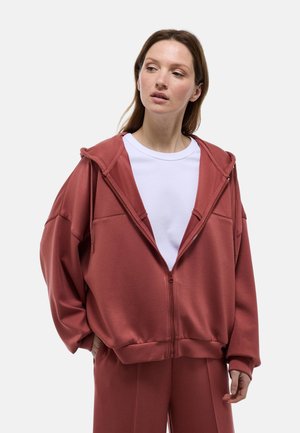 Haut de sweatshirt à zip de couleur rouille avec une capuche, des épaules tombantes et de grandes poches avant, fabriqué en tissu doux et texturé. Porté sur une chemise blanche.