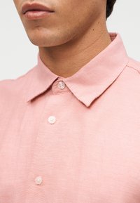 Άνδρας που φοράει ροζ παστέλ πουκάμισο με κλασικό γιακά, εμφανίζοντας το κάτω ήμισυ του προσώπου και τον λαιμό του πάνω σε απλό φόντο.