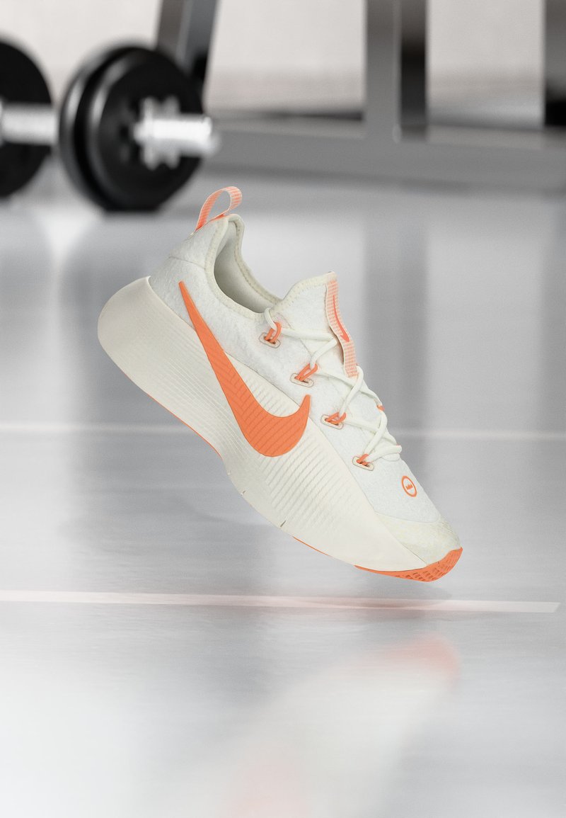 Λευκό και πορτοκαλί αθλητικό παπούτσι Nike που αιωρείται πάνω από έναν αντανακλαστικό πάτωμα γυμναστηρίου, με εξοπλισμό άρσης βαρών θολωμένο στο φόντο.