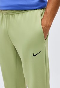 Άτομο που φορά ανοιχτοπράσινο παντελόνι Nike με ελαστική μέση και μαύρο λογότυπο Nike στο μηρό, χέρι στην τσέπη.