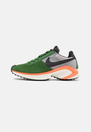 Low-top sneaker met groene suède voorkant, grijze mesh-zijden, zwart Nike-logo en witte zool met oranje accent op een effen achtergrond.