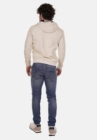 Felpa beige con cappuccio, polsini e orlo a coste; abbinata a jeans blu scuro. Progettata con una vestibilità comoda, vista posteriore visibile.