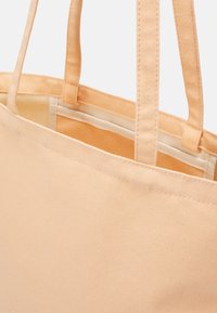 Sac fourre-tout en toile beige avec double bandoulière et coutures visibles le long des bords et de la doublure intérieure.