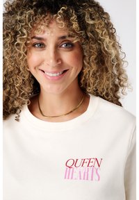 Crèmekleurige sweatshirt met de tekst "QUEEN HEARTS" in het rood en roze. Heeft een ronde halslijn en ribgebreide manchetten; eenvoudige, zachte katoenen textuur.