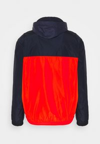 Huppari, jonka yläosa on tummansininen ja alaosa kirkkaan oranssi. Valmistettu sileästä, kevyestä materiaalista, jossa on joustavat hihansuut.