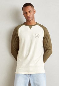 Pikad varrukad raglan-särk, millel on kreemivärvi keha ja oliivrohelised varrukad. Omab ümmargust kaelust ja logo trükki rinnal. Pehme kangast tekstuur.