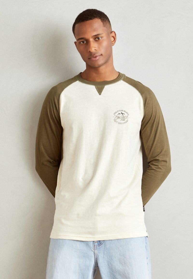 Pikad varrukad raglan-särk, millel on kreemivärvi keha ja oliivrohelised varrukad. Omab ümmargust kaelust ja logo trükki rinnal. Pehme kangast tekstuur.