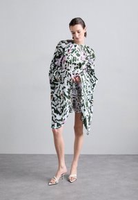 Blumiges Kleid in Grün, Weiß und Pink, mit weiten Ärmeln und lockerer Passform. Getragen mit weißen High-Heels.