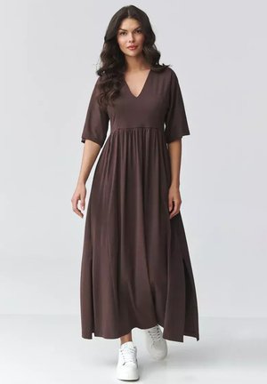 Kobieta o długich ciemnych włosach, ubrana w brązową, długą sukienkę maxi z dekoltem w serek i rękawami do łokcia oraz białe trampki, stojąca na tle jasnego, jednolitego tła.