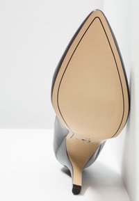 Chaussure à talon haut en cuir noir à bout pointu vue de la semelle, montrant une semelle beige et un talon aiguille fin sur fond blanc.