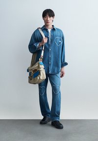 Young person wearing matching denim shirt and jeans with heart-shaped patches, carrying beige and blue shoulder bag, standing on gray floor.