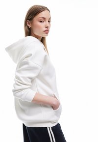 Giovane donna con un hoodie bianco, mani nella tasca frontale, che guarda di lato, con capelli lisci e orecchini a cerchio dorati.