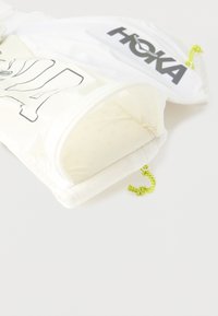 Λευκές υφασμάτινες θήκες με μαύρο λογότυπο «HOKA» και μαύρο περίγραμμα σχεδίου, η καθεμία με κίτρινο-νεον κορδόνι σύσφιξης σε λευκό φόντο.