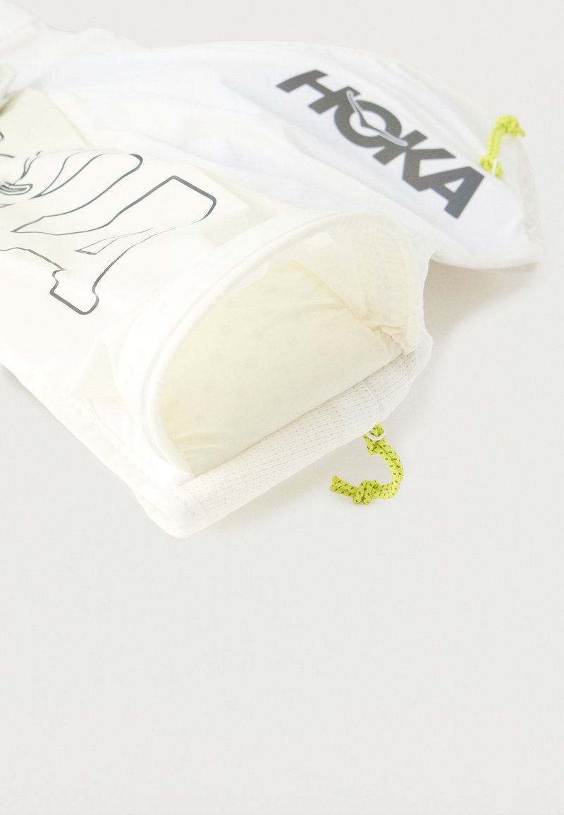 Sacchetti di tessuto bianco con logo "HOKA" nero e disegno contornato nero, ciascuno con chiusura a coulisse giallo neon su sfondo bianco.
