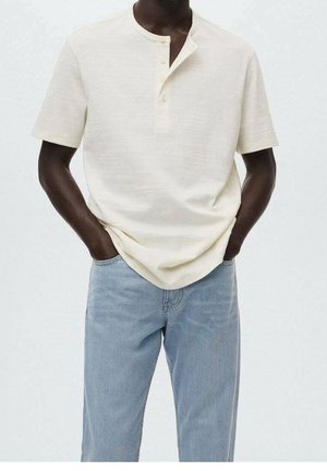 Dunkelhäutige Person, die ein off-weißes Kurzarm-Henley-Shirt und hellblaue Jeans trägt, mit den Händen in den Taschen vor schlichtem Hintergrund.