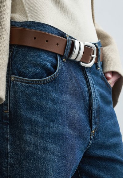 Καφέ δερμάτινη ζώνη με ασημένια αγκράφα, φορεμένη πάνω από μπλε τζιν. Το τζιν έχει ξεθωριασμένη υφή και εμφανείς λεπτομέρειες ραφής.