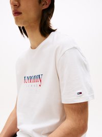 T-shirt en coton blanc avec "tommy" en rouge et "JEANS" en bleu. Présente un petit logo sur la manche gauche, des manches courtes et un design col rond.