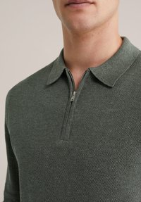 Temno zelena polo majica z zadrgo, teksturiranega tkanja, z klasičnim ovratnikom in srebrnim detajlom zadrge na vratu.